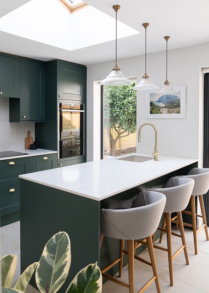 Green shaker kitchen with a white quartz peninsula, brass tap and three upholstered bar stools